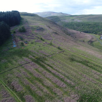 A photo showing the site within the Breamish Valley. In the background next to the blue plastic is the trench. Medieval ridge and furrow remains dominate the foreground.